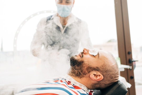 Crop anonymous beauty master in mask near adult bearded male client with cotton pads on closed eyes during vapor procedure in barbershop