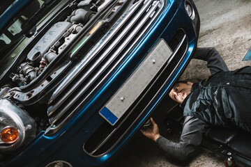 Young focused auto service man checking up vehicle with open bonnet while lying on mat in workshop