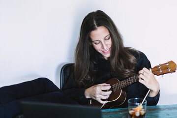 Happy young female looking at laptop screen and playing ukulele while having online lesson at shabby wooden table at home