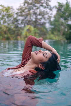 Side view of tranquil young ethnic Indian female in suit touching hair while relaxing in clean water of tropical lake in nature