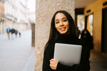 Charming young ethnic female with netbook smiling at camera while standing at stone column on city street