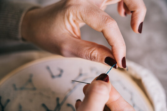 Crop anonymous female with manicured hands threading needle while sitting at table with threads and hoop and doing embroidery