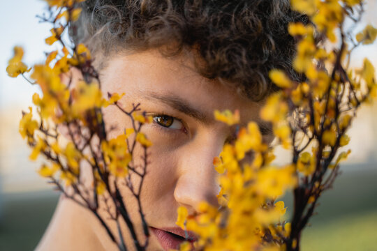 Crop male covering face with bright blossoming flower sprigs while looking at camera in daylight