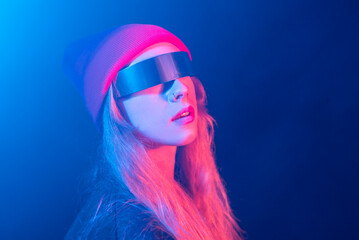 Trendy young female millennial with long blond hair in futuristic sunglasses adjusting hat while standing in dark room with neon illumination