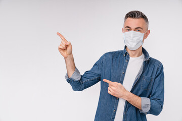 Cheerful middle-aged man in medical mask against Covid 19 pointing at copy space isolated over white background