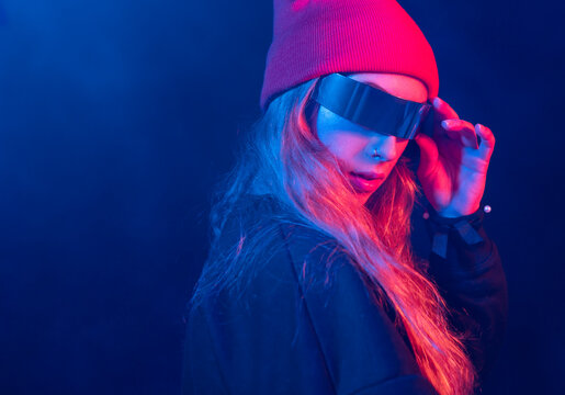 Trendy young female millennial with long blond hair in futuristic sunglasses adjusting hat while standing in dark room with neon illumination