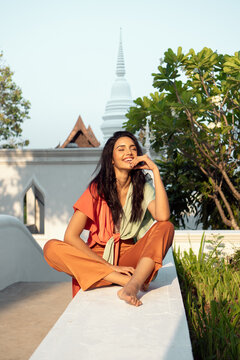 Barefoot young happy ethnic female in stylish colorful outfit with legs crossed with eyes closed while sitting on border on sunny terrace