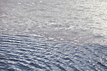 Shiny silver melting ice surface and water. Sparkling ice on clear water surface close-up. Melting chunks of ice in dark blue water. White and transparent ice float in the water. Spring ice melting. 