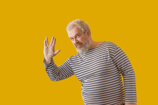 A man with a gray beard, gray hair, wearing a sailor's shirt, shows the "vulcan salute" sign. On a yellow isolated background.