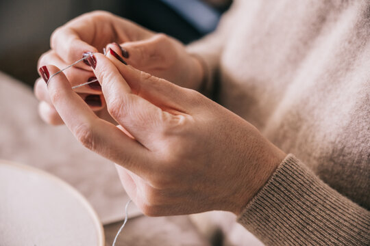Crop anonymous female in casual outfit threading needle for sewing work while sitting at table in light studio