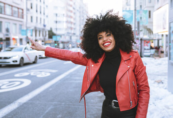 Full body of positive young African American female wearing trendy outfit standing on snowy road and catching taxi