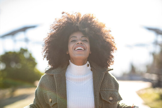Cheerful young African American female with curly hair in stylish warm outfit smiling and looking at camera while resting in park at lakeside on sunny autumn day