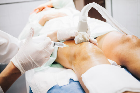Detail of a hand with white latex gloves pricking a patient's knee with a needle while performing an ultrasound test