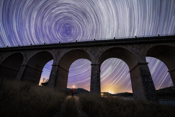 From below of traveler with flashlight standing under stone viaduct bridge and night sky with stars in long exposure