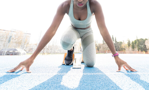 From below of African American female runner in starting blocks sitting in crouch position while getting ready for sprint at stadium during workout