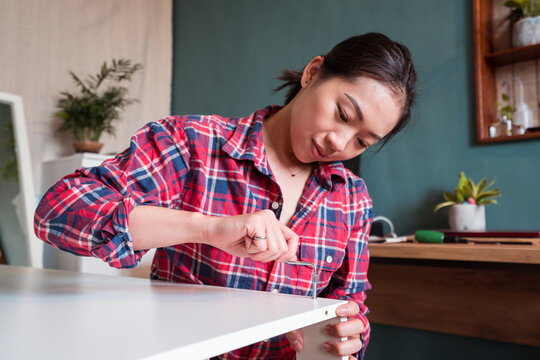 Ethnic Asian female sitting on floor and screwing wooden board while assembling new furniture at home