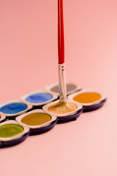 Crop unrecognizable creative person dipping paintbrush on colorful palette of watercolor paints against pink background