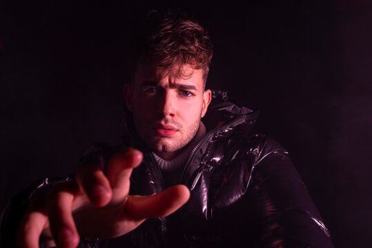 Thoughtful young unshaven guy in trendy outfit reaching out hand towards camera against black background
