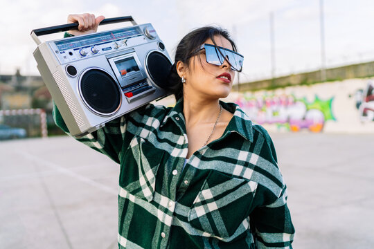 Young trendy female in sunglasses reflecting cloudy sky listening to song from old tape recorder against graffiti wall in sunlight