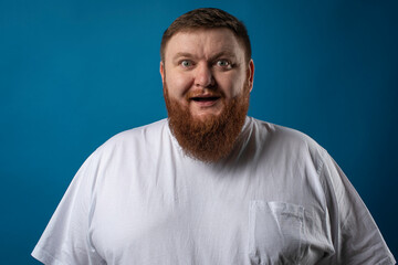 Fototapeta premium portrait of a bearded cheerful, emotional and interesting man in a white T-shirt on a blue background, showing various hand gestures. Cheerful guy show business artist, host of a glamorous show. Body 