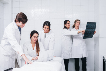 A group of doctors working on a laptop while two female doctors examine an X-ray in a white hospital room