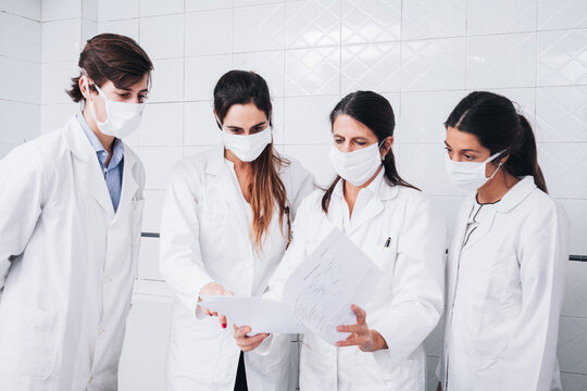 A group of doctors wearing a face mask examining medical reports in a hospital room and dressed in white coats