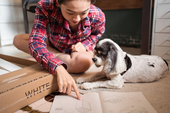 Smiling Asian female sitting on floor with fluffy dog and reading instruction while assembling furniture in new apartment