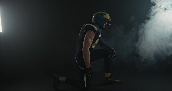 Side view of male American football player in helmet and uniform kneeling in dark studio with steam and light