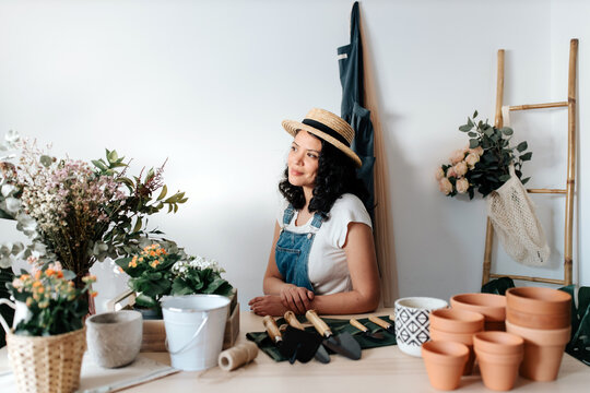 Young female horticulturist in straw hat sitting near flowers on table with assorted tools at home