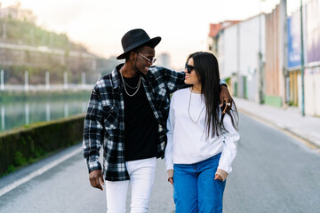 Young content multiracial partners in trendy clothes and sunglasses talking while looking at each other and strolling on city roadway