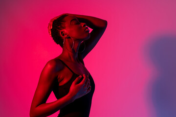 Side view of self assured young African American female model in black top touching face with closed eyes while standing against pink background in neon light