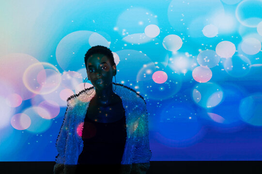 African American woman in trendy fur coat standing under vivid blue projection with specks of light