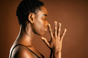 Side view of African American female with trendy earrings with praying hands against brown background with eyes closed