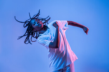 Dynamic African American teenage girl making movement while performing urban dance in neon light against blue background