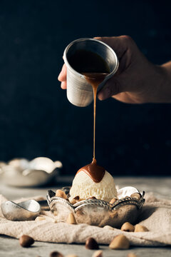 Unrecognizable crop person pouring sweet caramel topping on vanilla ice cream scoop in bowl placed on table with scattered nuts