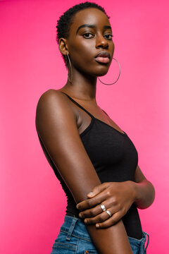 Confident young fit African American female model in black top and jeans looking at camera while standing against pink background