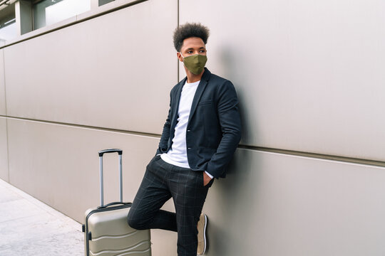 African American male tourist in protective mask standing near suitcase and leaning on airport building while waiting for flight during coronavirus epidemic