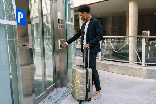 Side view of African American male tourist with suitcase and in protective mask pressing button of elevator in airport while traveling during coronavirus pandemic