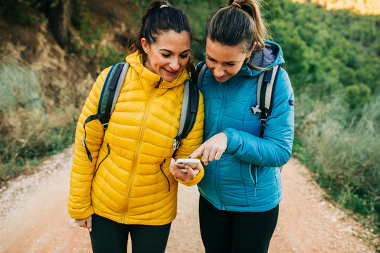Cheerful female hikers with backpacks and in warm jackers standing on road in nature and orientating on GPS map on smartphone during trekking