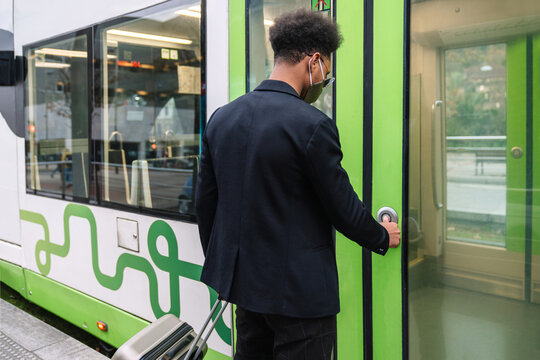 Back view of African American male tourist in mask and with suitcase pressing button and entering modern train while standing on platform during coronavirus