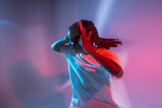 Serious African American teenage girl in headphones dancing in studio with bright glowing neon lights