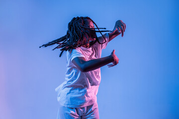 Dynamic African American teenage girl making movement while performing urban dance in neon light against blue background