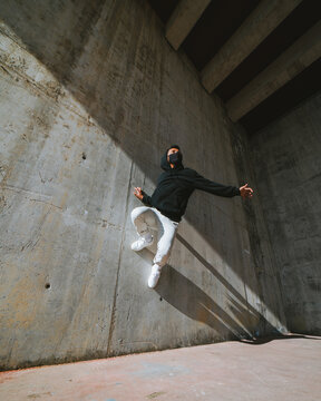 From below of unrecognizable young male in trendy outfit and mask performing trick on concrete wall while dancing in abandoned building on sunny day