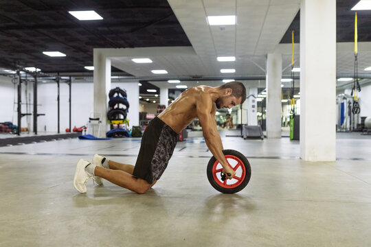 Side view of strong young muscular sweated young guy using wheel roller while doing abs exercise in contemporary sports club