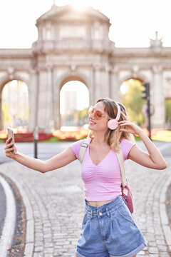Cheerful teenage female in casual outfit and sunglasses taking self portrait while listening to music on Madrid street