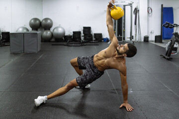 Powerful muscular young male athlete with naked torso standing in Side Plank and lifting heavy kettlebell during workout in gym