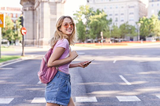 Side view of young woman walking on pedestrian crossing and checking route on mobile phone in Madrid