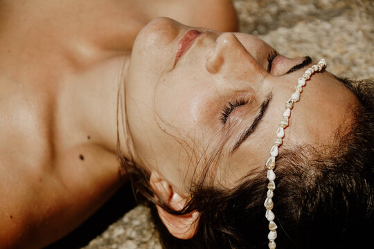 From above of crop peaceful young female sunbathing with closed eyes while lying on sandy beach with seashell beads on face