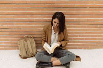 Positive female student sitting on floor and reading book while studying in university corridor