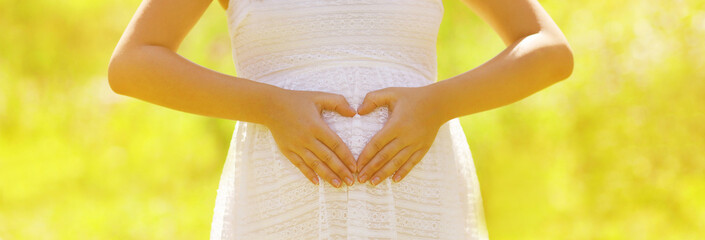 Fototapeta na wymiar Close up of pregnant woman showing heart shaped hands on belly in sunny summer day
